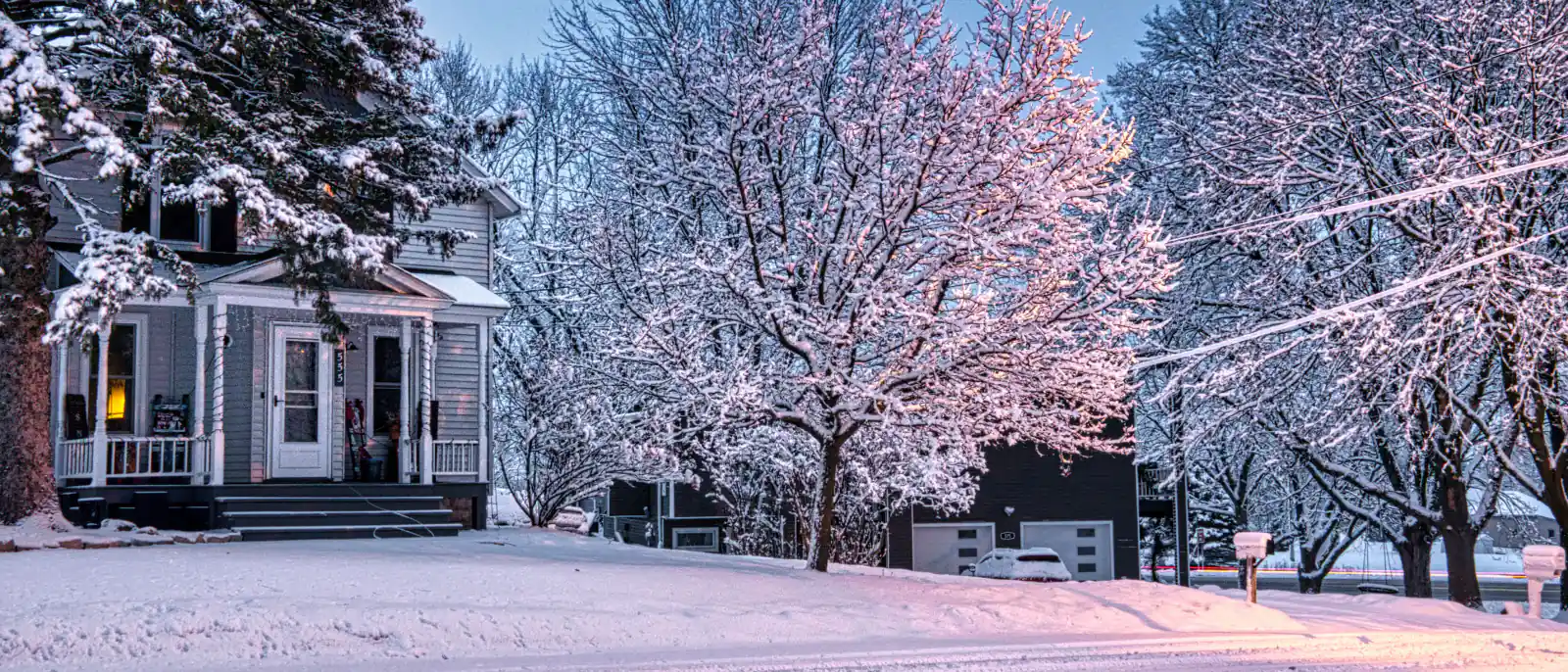 Older two-story frame house on a snow-packed residential street at dusk, snow load on the bare trees and drifts across the yard