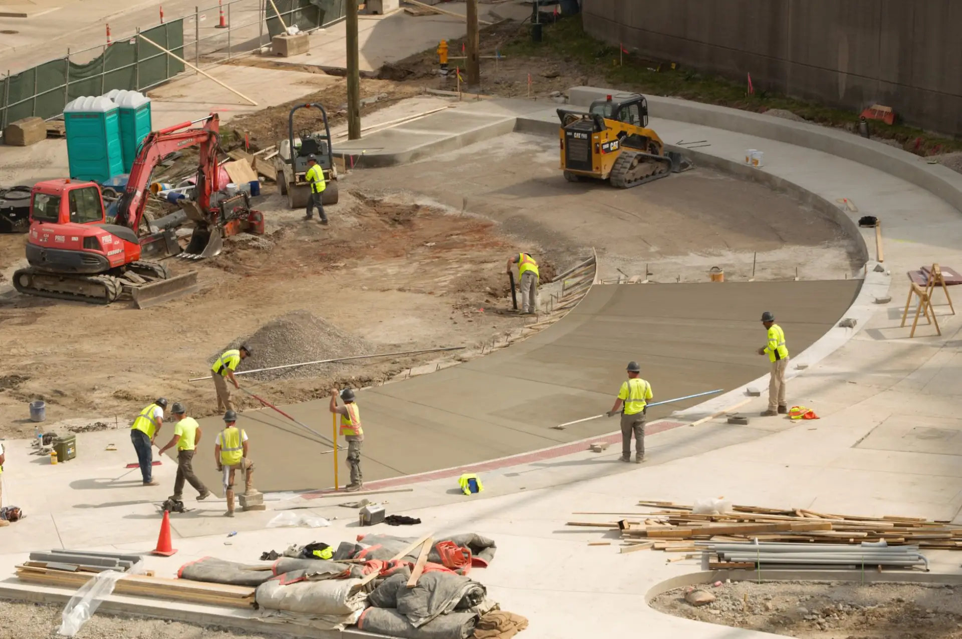 Commercial concrete flatwork being placed and finished by a crew, with an excavator and skid steer working the site