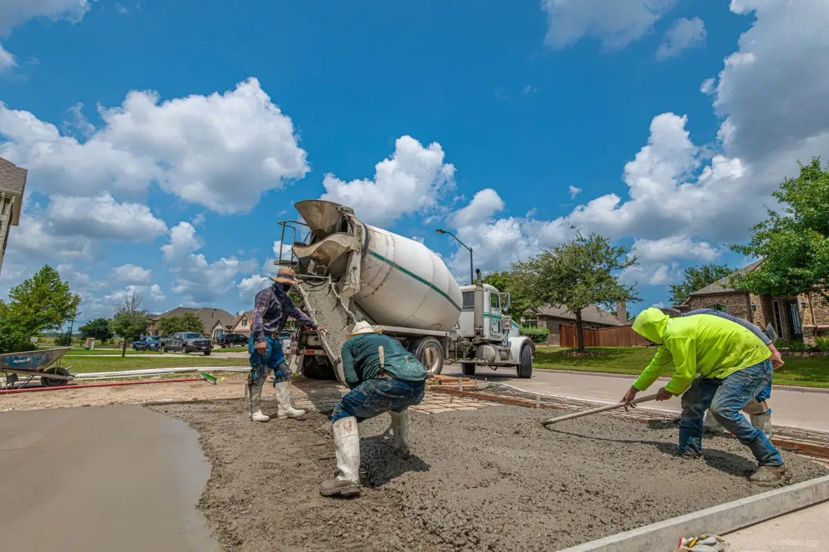 Ready-mix truck on a residential street while two workers in high-visibility gear screed a fresh concrete driveway
